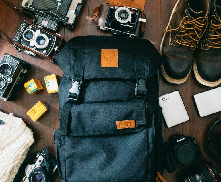 high-angle photo of black bag beside cameras, portable HDD and black leather lace-up boots